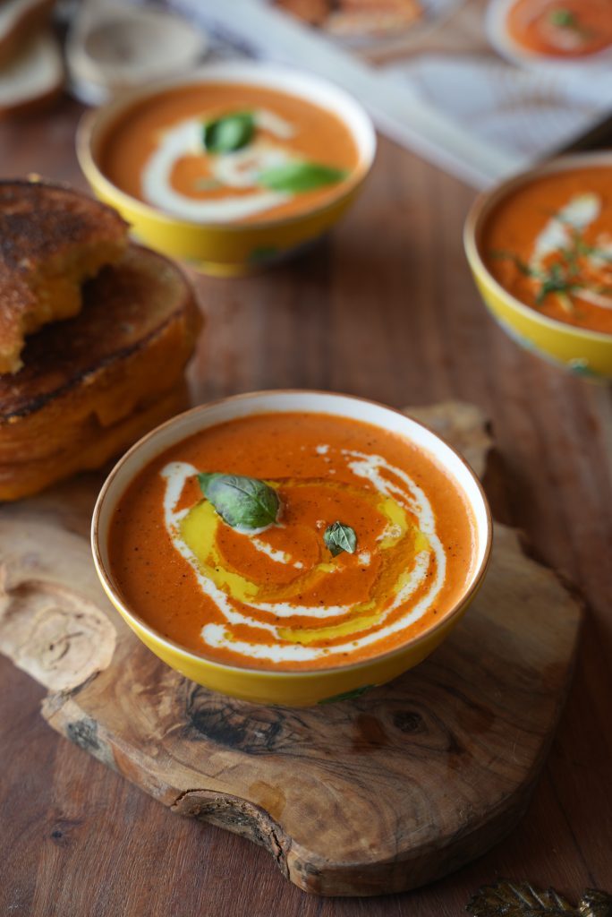A bowl of creamy tomato soup with a cream swirl, basil and grilled cheese on the side